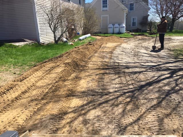 A man is standing on a dirt road next to a house holding a shovel.