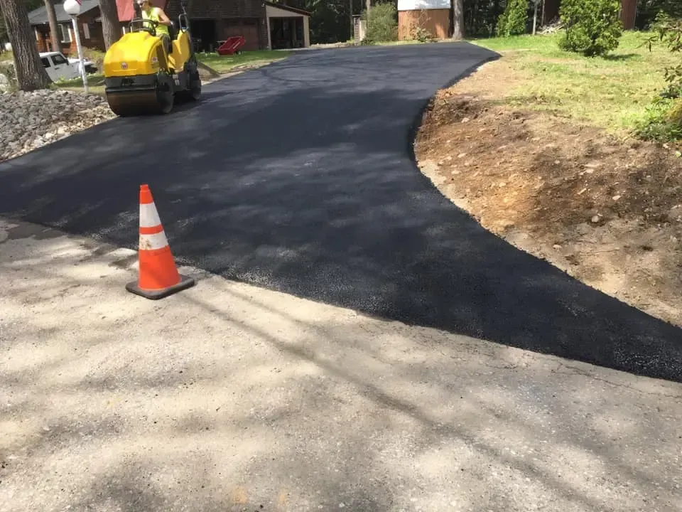 A yellow roller is laying asphalt on a driveway next to a traffic cone.