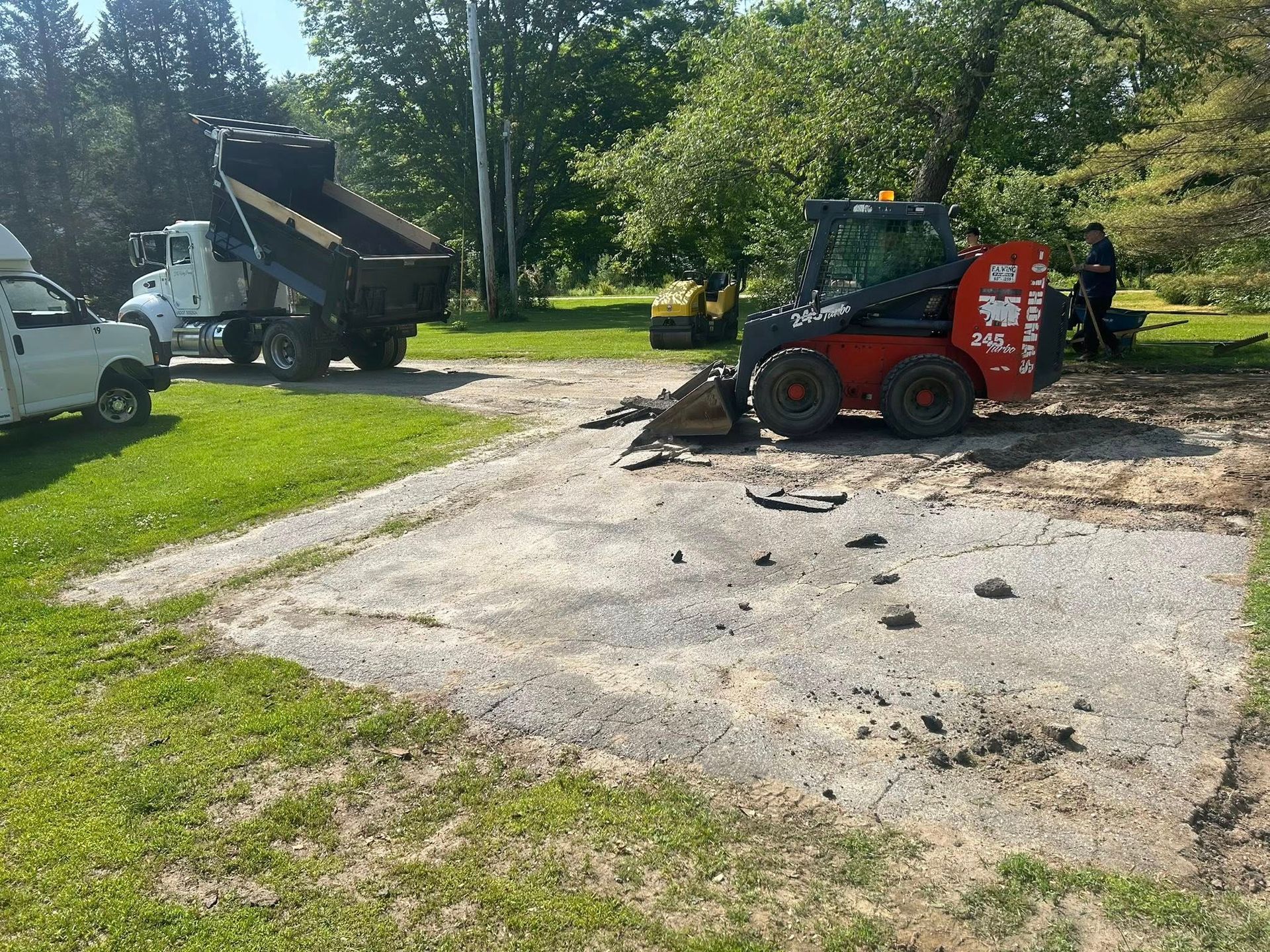 A red skid steer is parked on the side of a dirt road next to a dump truck.