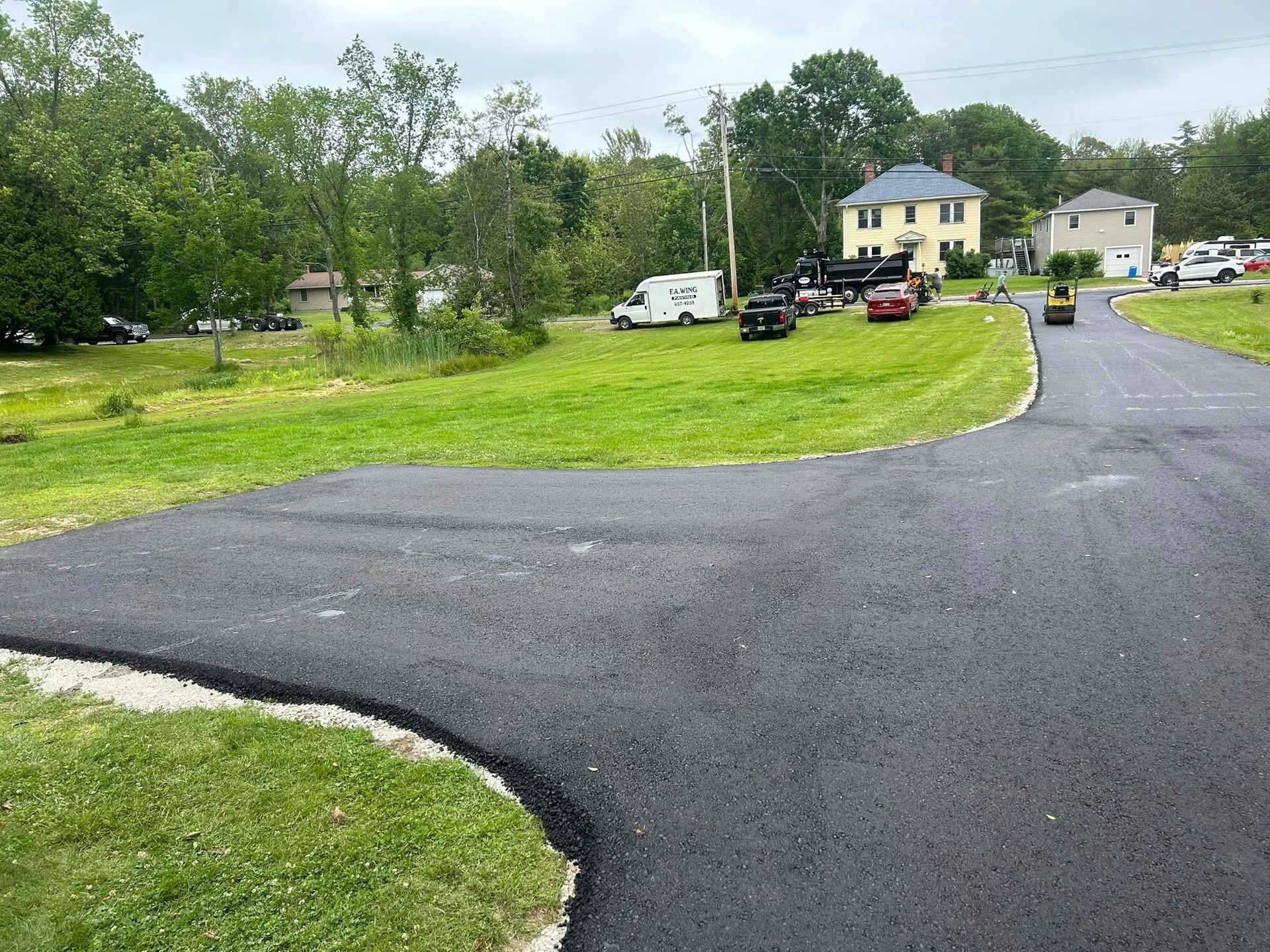 A black road going through a grassy field with a house in the background.