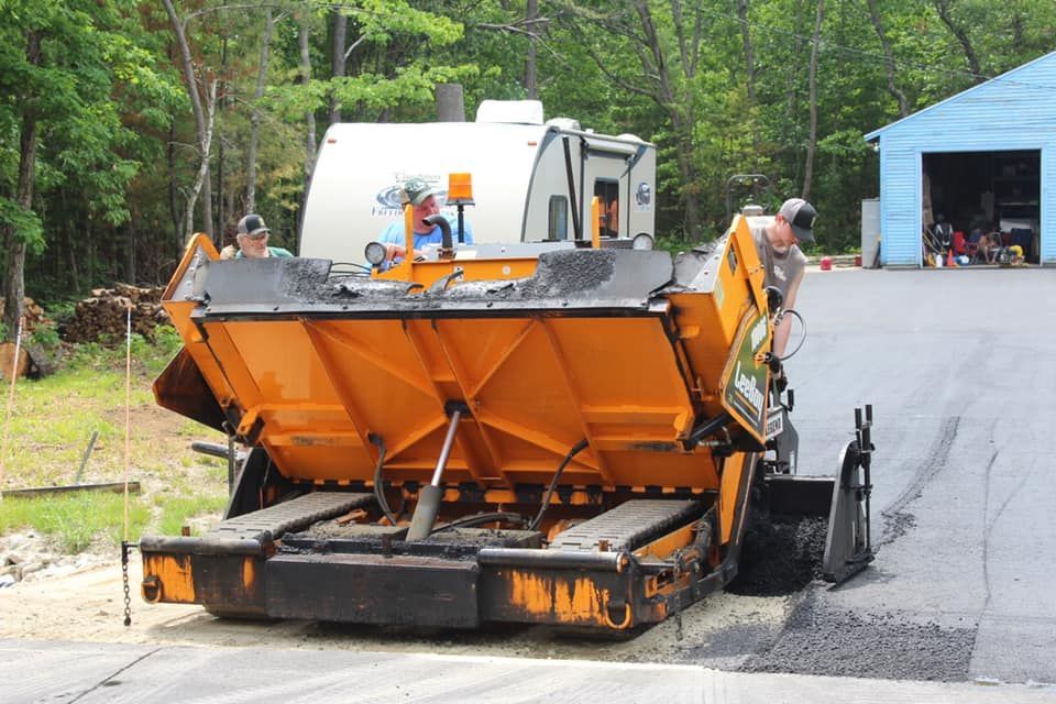 A yellow and black asphalt paving machine is parked in front of a blue garage.