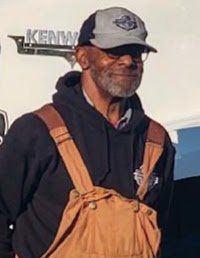 A man wearing overalls and a hat is standing in front of a truck.