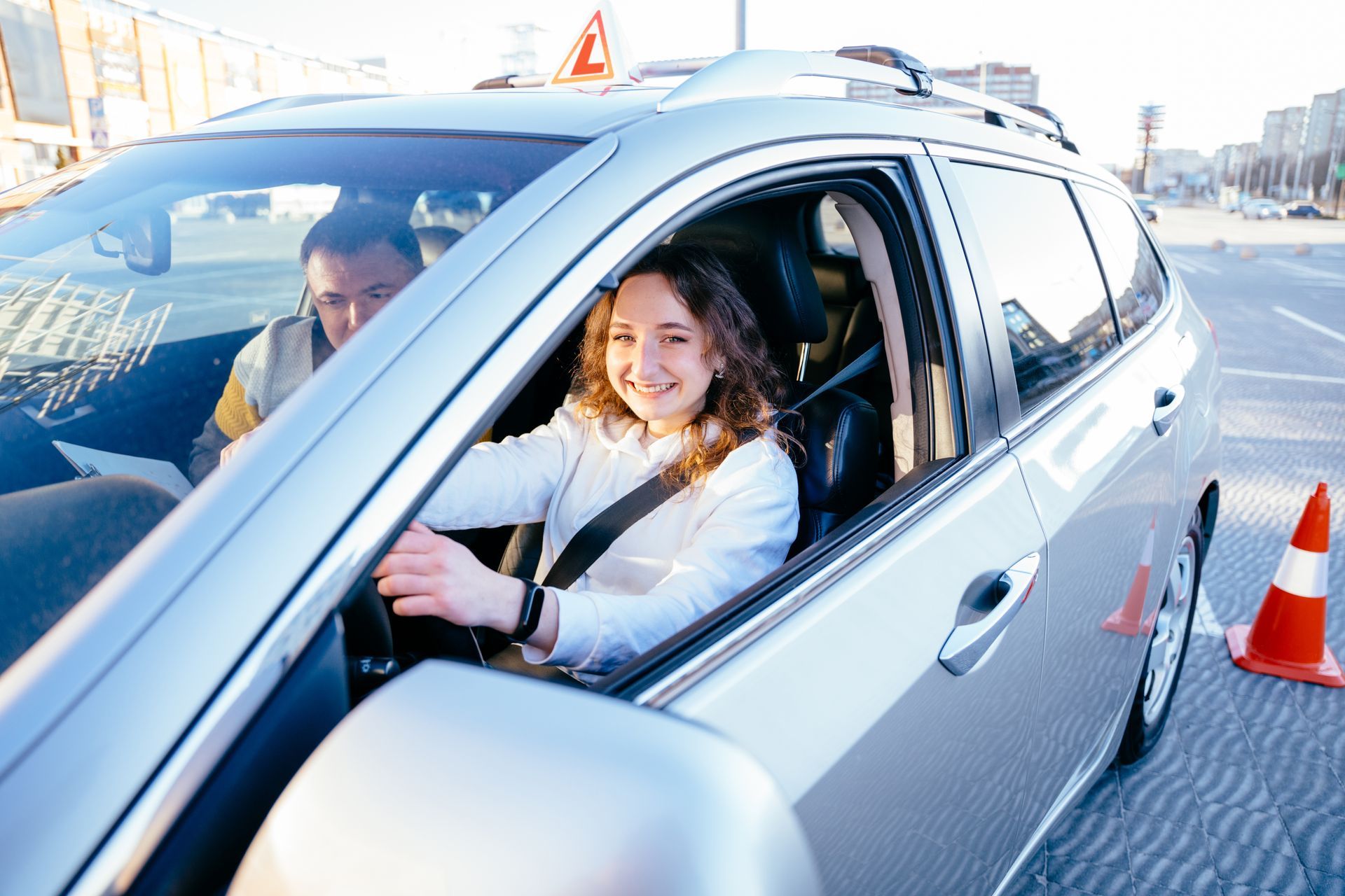 Young woman smiles driving a car during a driving lesson. Instructor in passenger seat. Orange traffic cone.