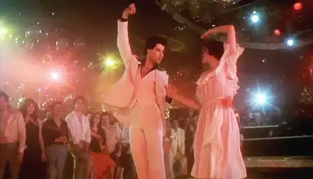 Two dancers in a disco, John Travolta in white suit, woman in pink dress, crowd watching.
