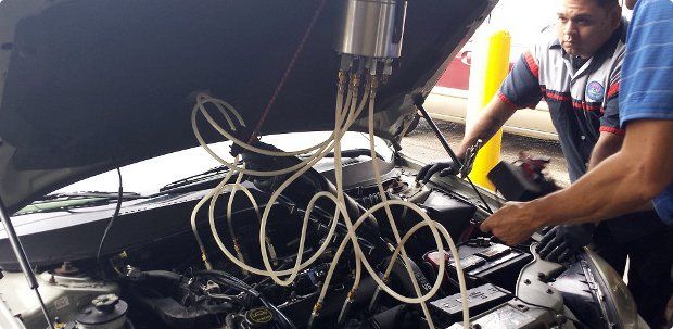 Mechanic working under a car hood with tangled cables and tools in a garage | AACCEL Emissions & Auto Repair Specialists