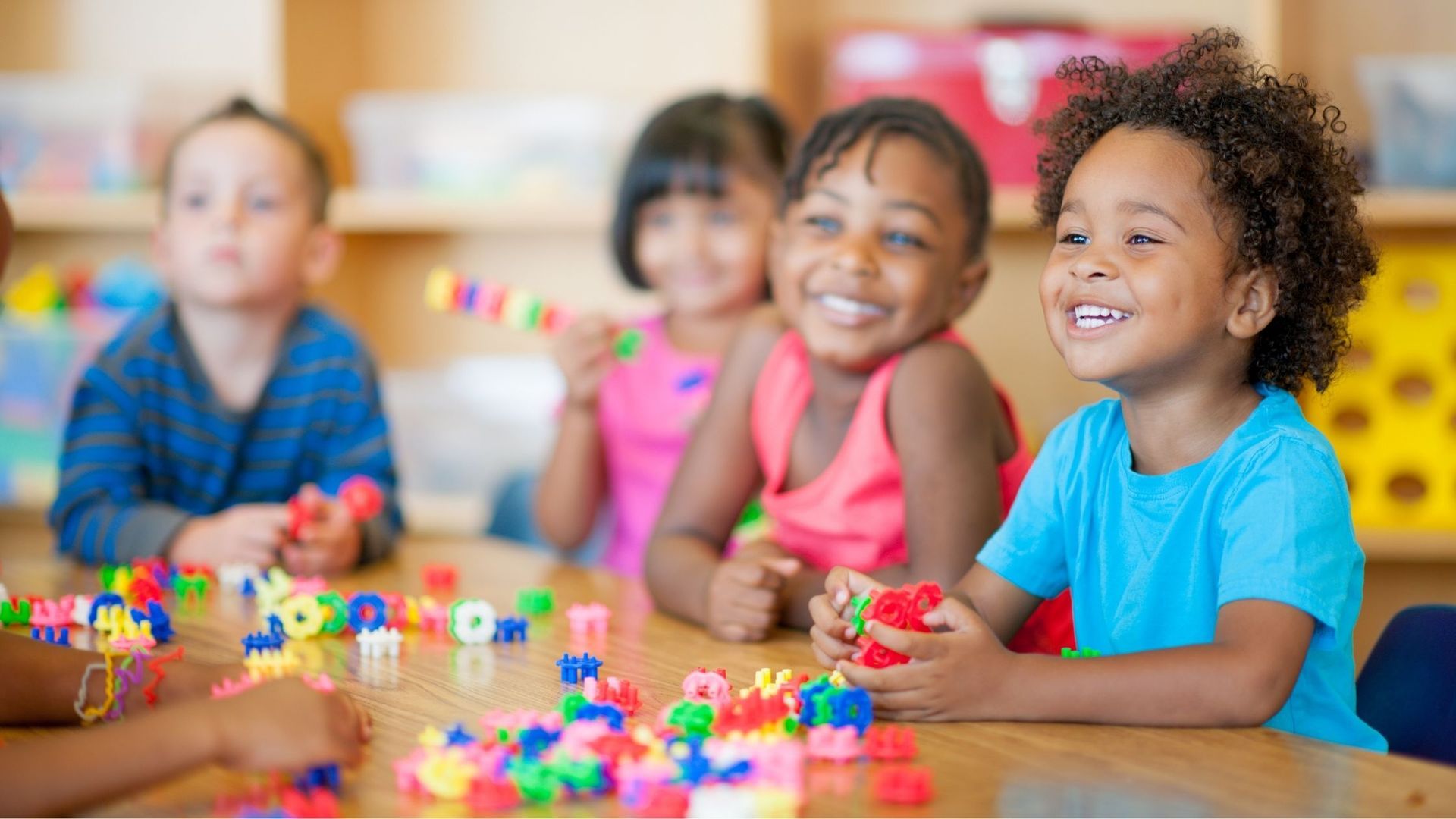 Children at a table smiling, playing with colorful building blocks in a bright classroom.