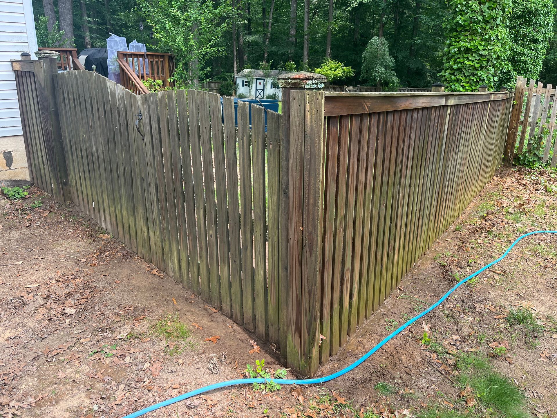 Weathered wooden fence with angled corner, surrounded by dirt and grass, a blue hose lies in front.