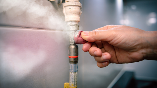Hand turning a red valve, steam escaping from a pipe against a metal surface.