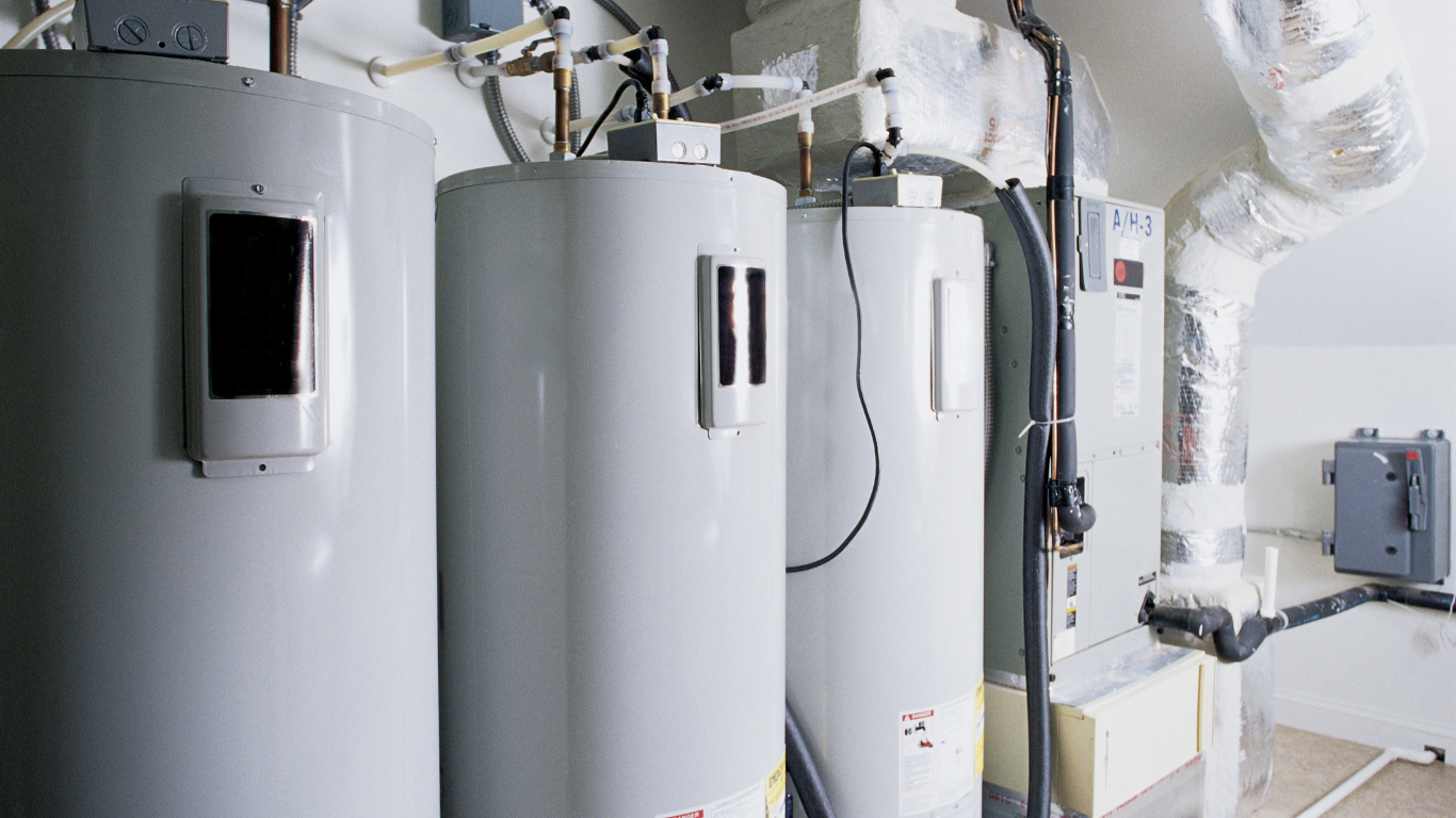 Three gray water heaters in a utility room, with pipes and ventilation visible.