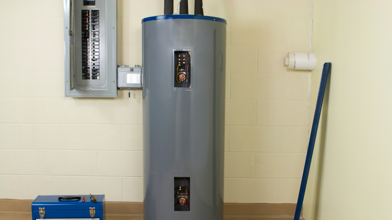 Electric water heater against a beige wall, near an electrical panel and a blue tool box.