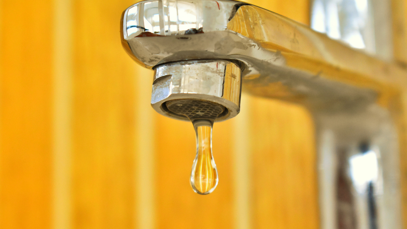 Dripping faucet, water droplet falling. Silver faucet against a blurred yellow background.