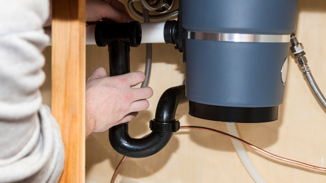 Hands adjusting black pipe under a kitchen sink near a garbage disposal unit.