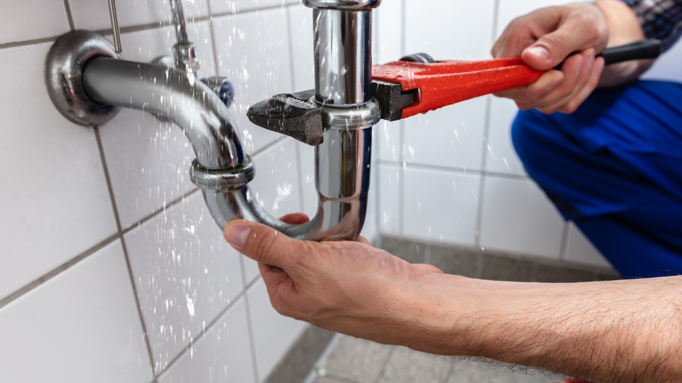 A plumber with a red wrench fixing a leaking sink pipe in a tiled bathroom.