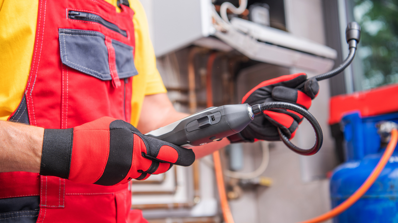 Person in red overalls, black and red gloves holding a black hose, near a boiler and propane tank.