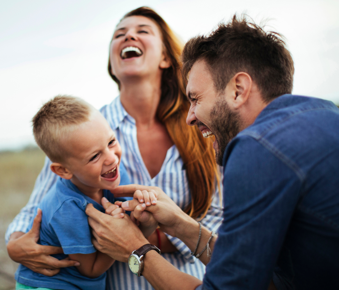 A joyful young child being held and tickled by two adults in an outdoor setting, all laughing together.