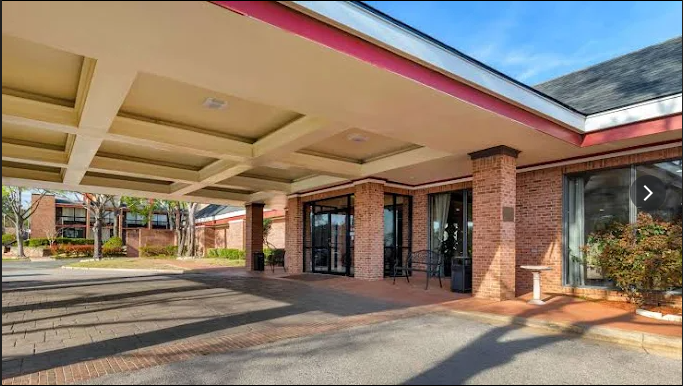 Covered brick building entrance with red trim and large overhang, seen from a driveway.
