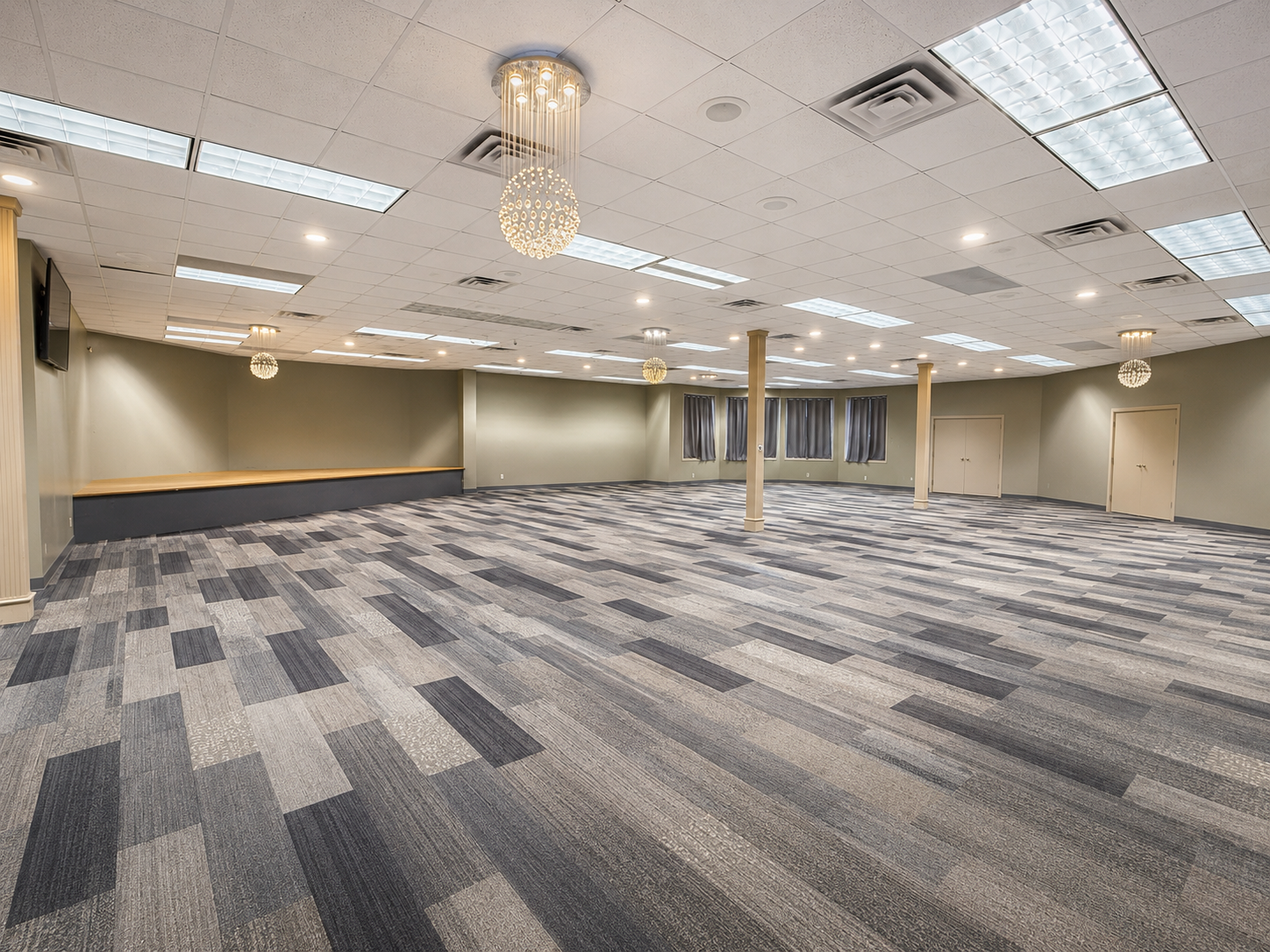 Empty banquet hall with gray patterned carpet, beige walls, chandeliers, and fluorescent ceiling lights