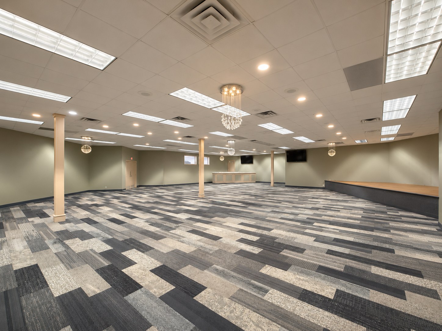 Empty carpeted conference room with fluorescent lights, beige walls, and a small stage at the back.