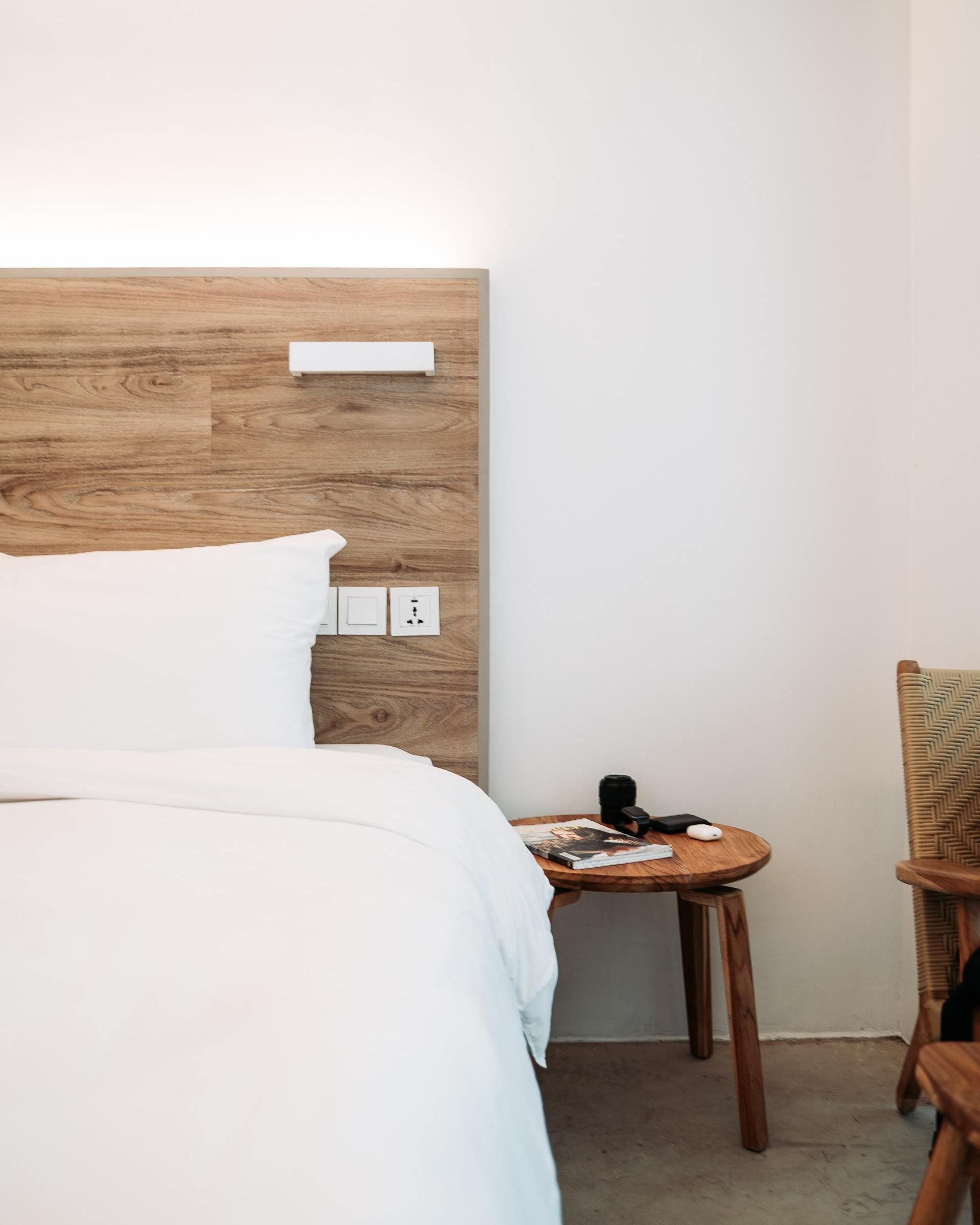 Minimal hotel room with bed, wooden headboard, and small side table by a blank wall.