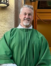 A man wearing glasses and a blue sweater is smiling in front of a stone wall.