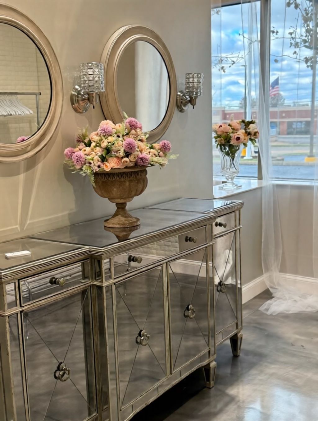 Mirrored credenza with floral arrangement, round mirrors, sconces, and flowers by a window.