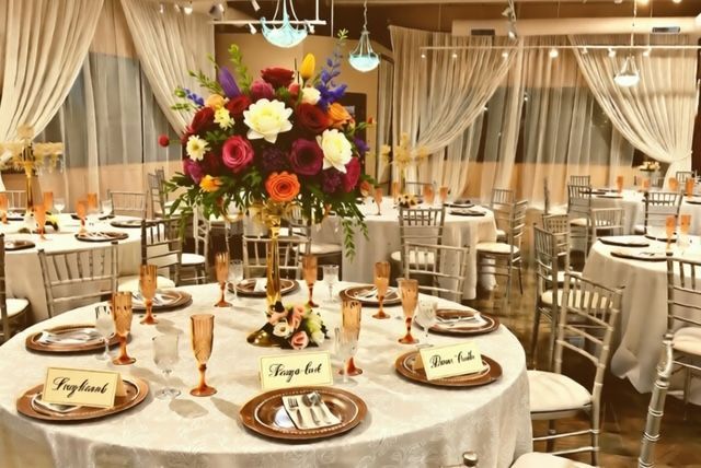 Elegant reception hall with round tables set for a wedding; large floral centerpiece.