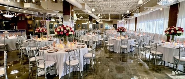 A spacious wedding reception hall with round tables, floral centerpieces, and silver chairs.