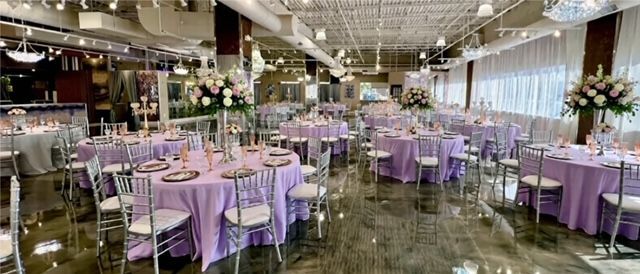 A large event space with round tables, lavender linens, silver chairs, and floral centerpieces.