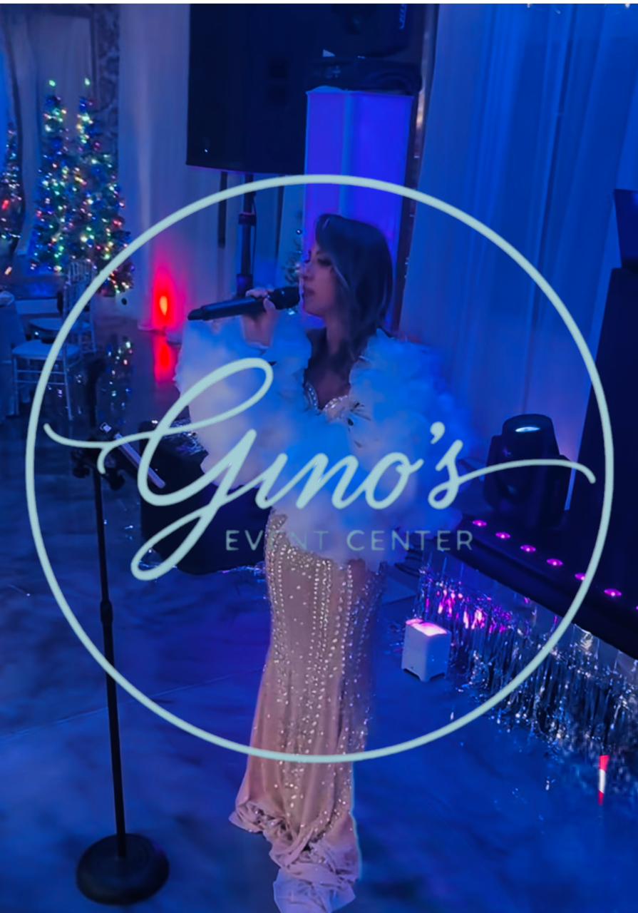 Woman singing on stage at Gino's Event Center. She wears a sparkly gown and a feather boa, with blue stage lighting.
