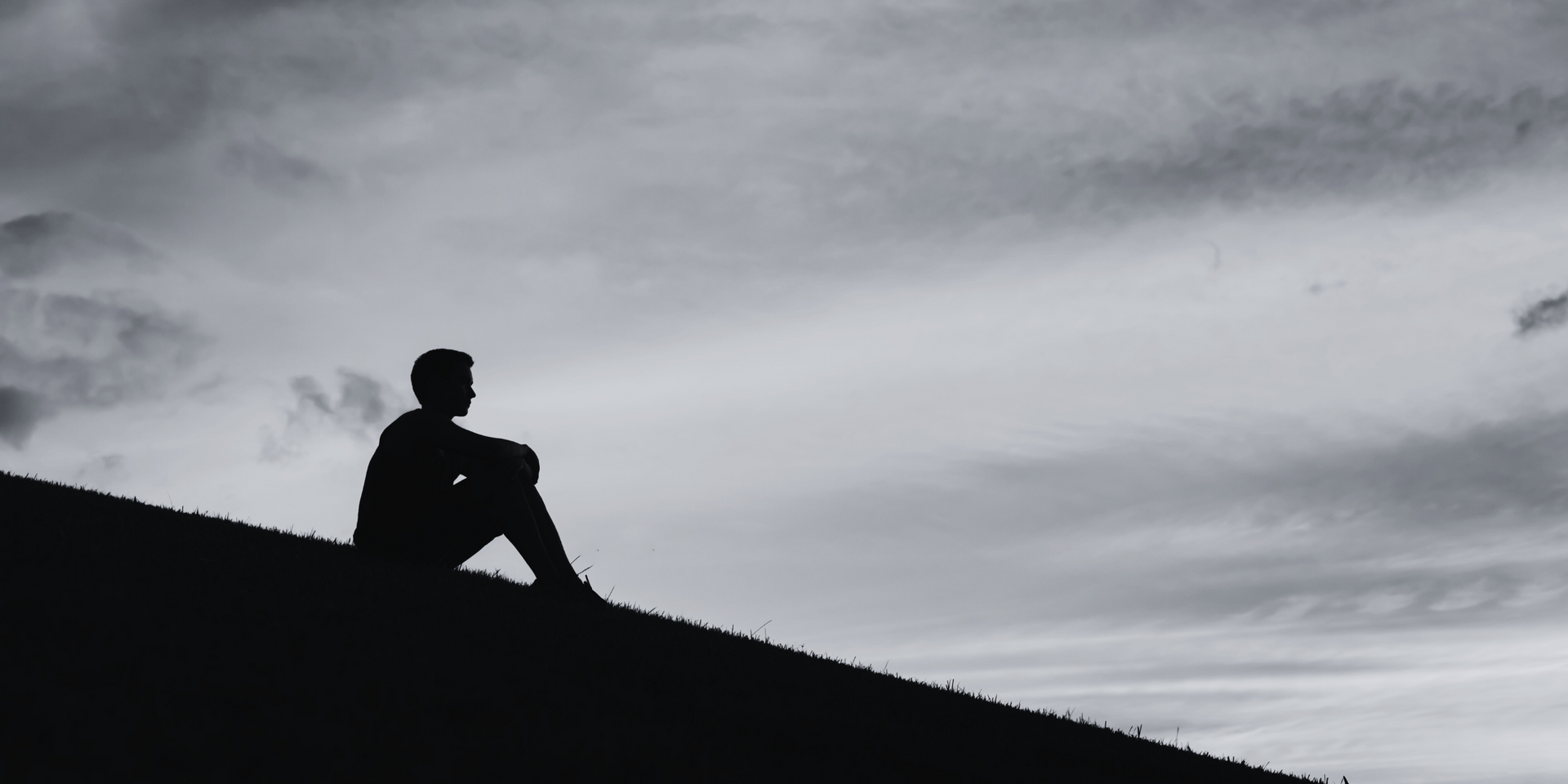 Silhouette of a person sitting on a grassy hill, gazing at a cloudy sky.