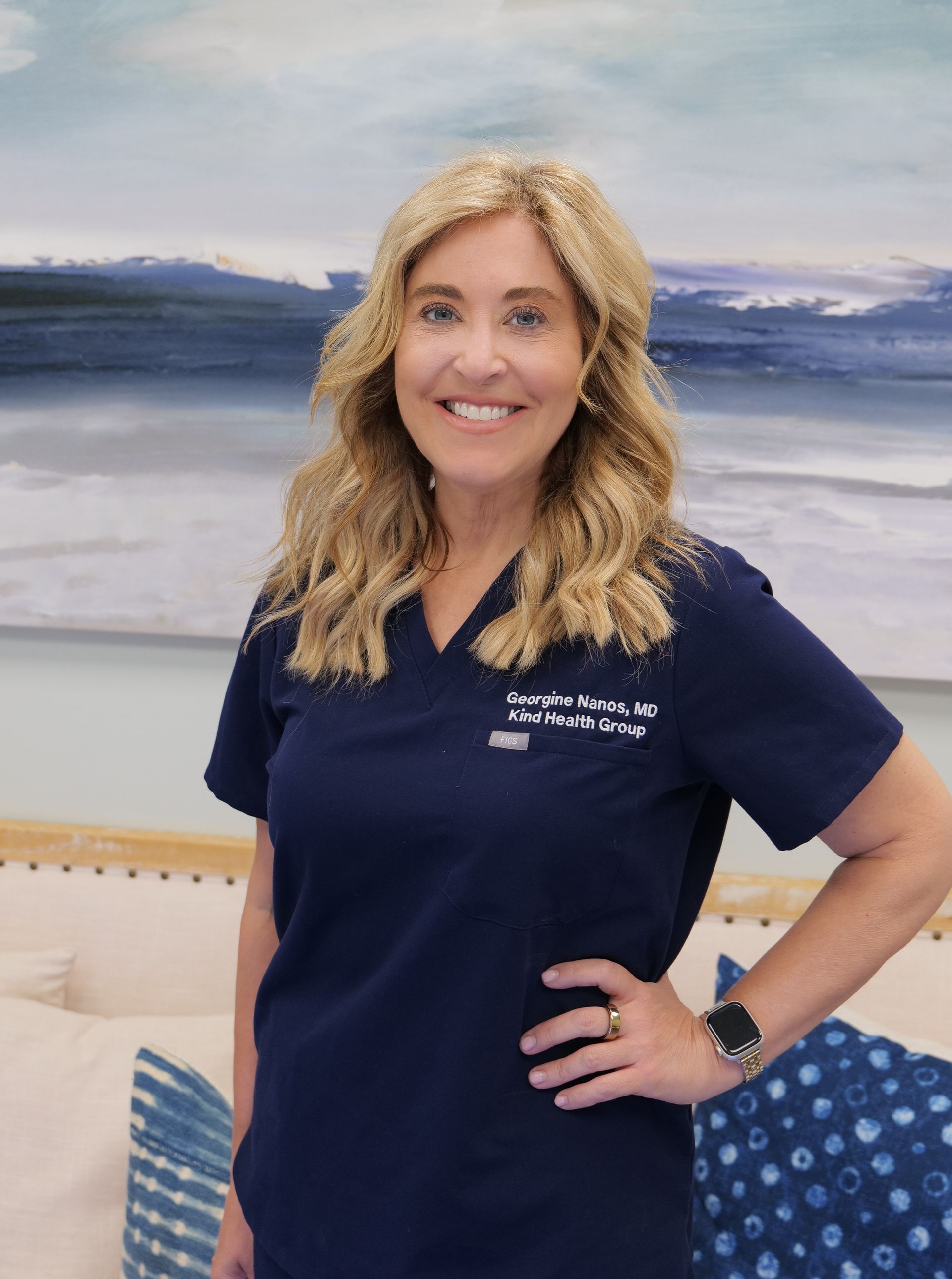 A smiling person with long blonde hair, wearing a navy blue medical scrub top, standing in a room with blue art.