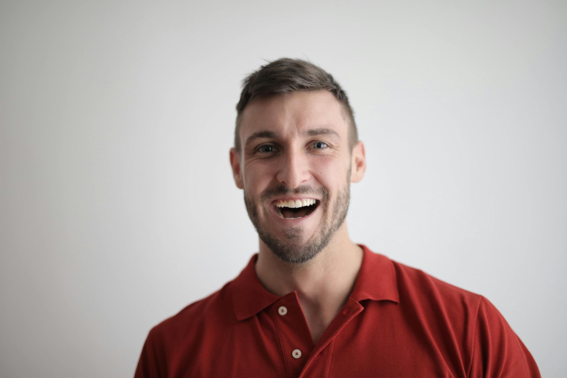 Man in red polo shirt smiles widely against a light gray background.