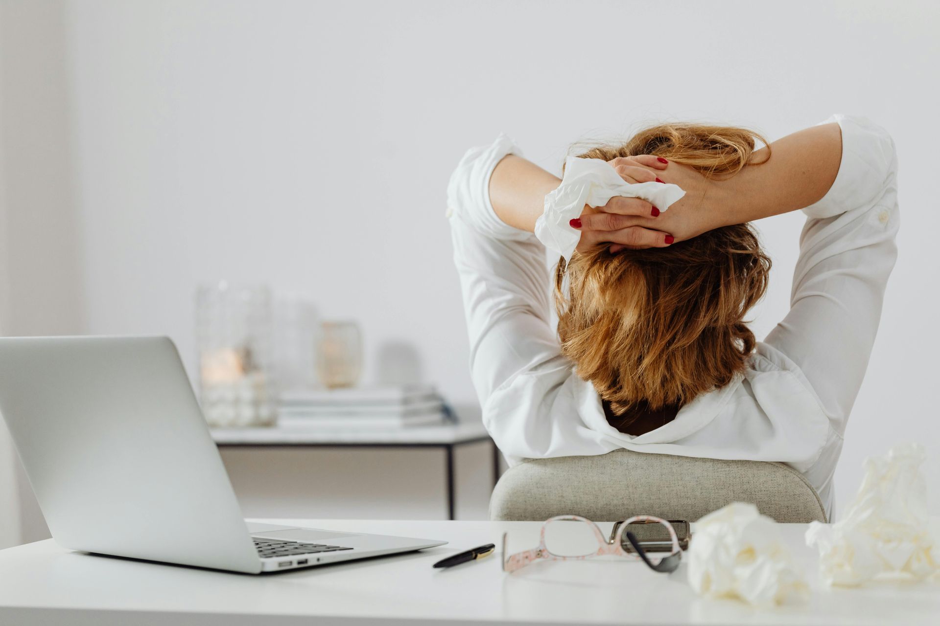 Person leaning back in an office chair with hands behind head beside a laptop and glasses on a desk