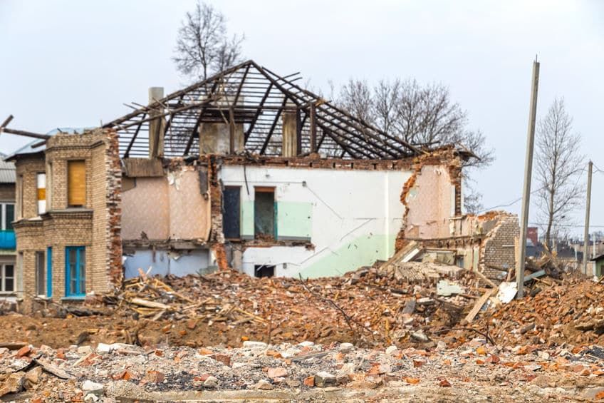 A partially demolished brick building with exposed interiors, rubble, and bare roof structure under a cloudy sky.