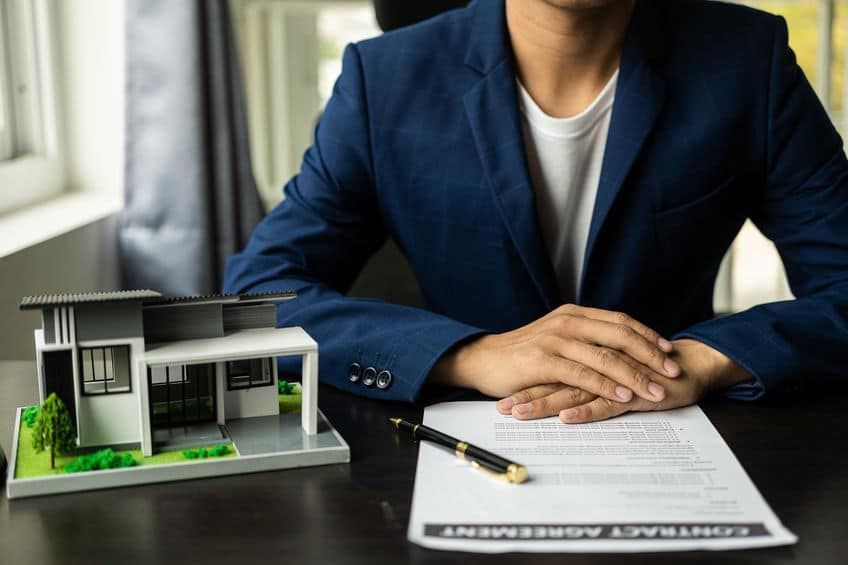 Person in blue blazer, hands folded, sits at a desk with a house model, contract, and pen.