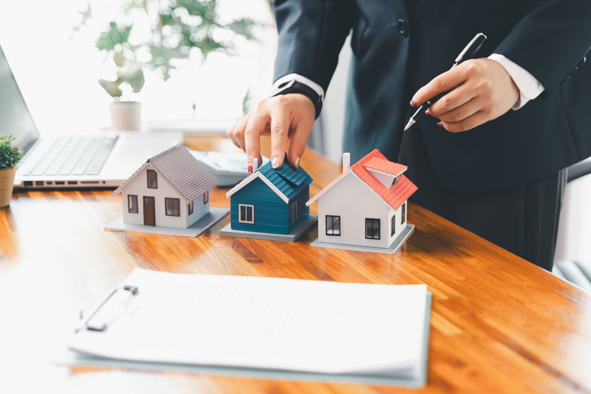 Person in suit pointing at blue house model among other miniature houses, paperwork and laptop on table.