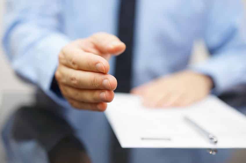Hand outstretched towards the viewer; person in blue shirt and tie; a document and pen are on a table.