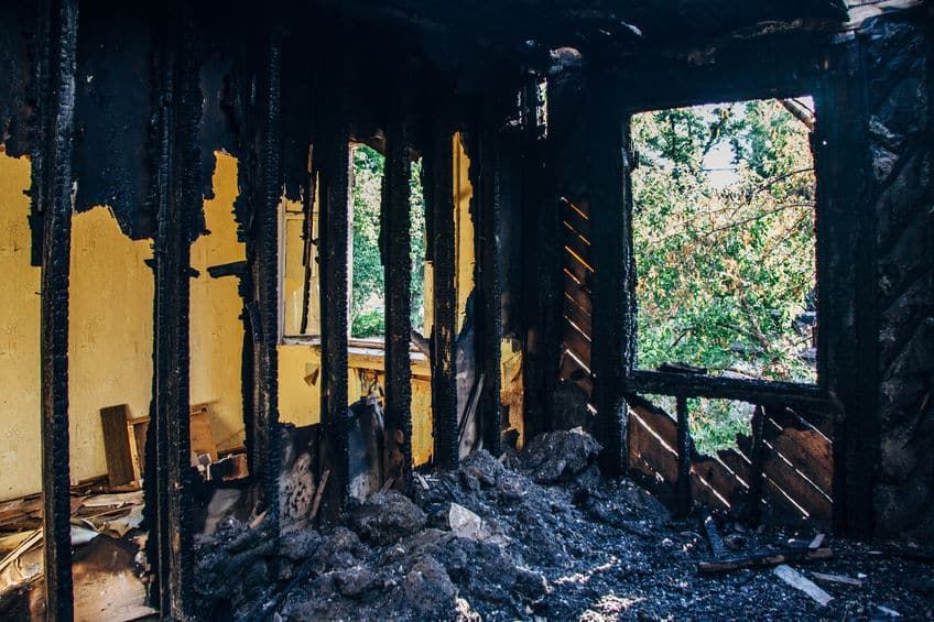 Interior view of a burnt room, charred walls and debris, a window shows green trees outside.