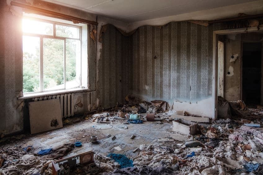 Dilapidated room with stripped wallpaper, debris on the floor, and sunlight through a window.