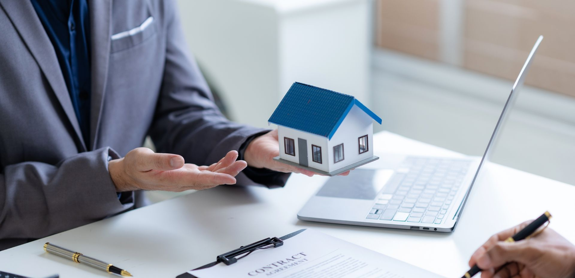 Person in a suit holding a model house, next to a laptop and a person signing a document.