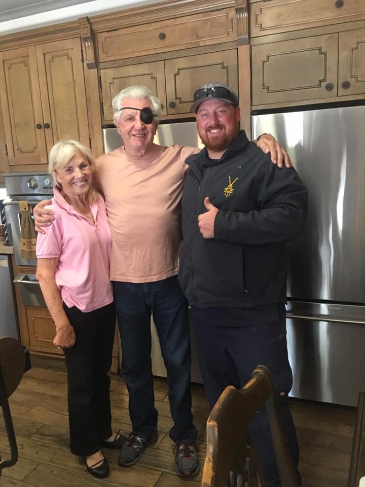 Three people in a kitchen pose together. Man with eye patch in center, woman on left, man on right giving thumbs up.