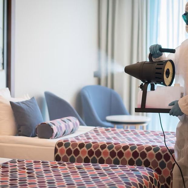 Person in protective suit sanitizing a hotel room with spray device.
