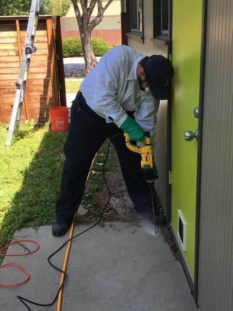 Person in work attire using a jackhammer on a concrete surface next to a building.