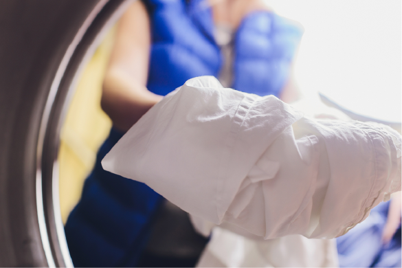 A person in a blue vest reaching into a clothes dryer to remove a piece of white laundry.