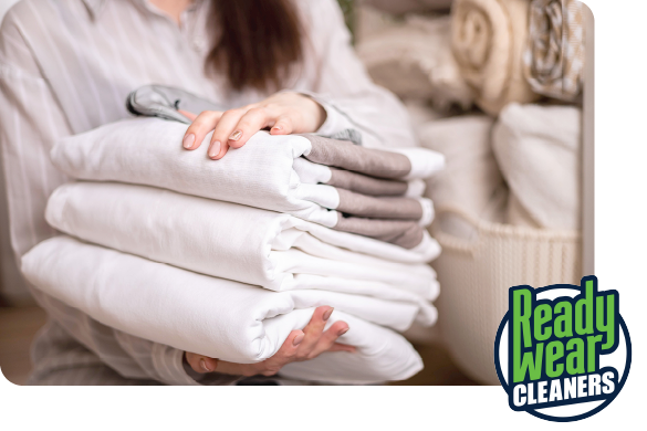 A person holding a stack of neatly folded white laundry with gray hangers, beside a basket of towels and brand logo.