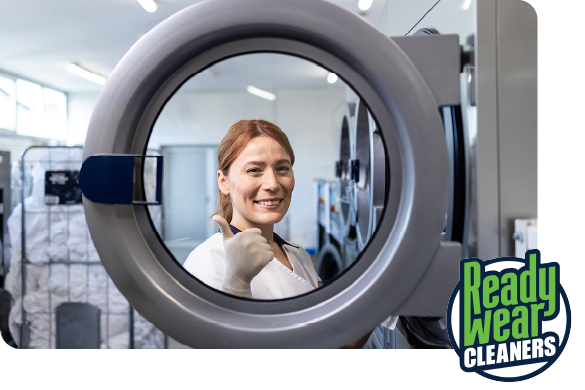 A person in white work clothes giving a thumbs-up through the circular door of an industrial laundry machine.