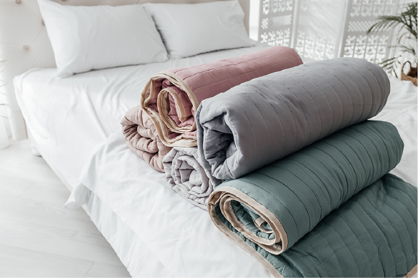 A stack of four rolled-up, quilted blankets in shades of pink, light gray, dark gray, and sage green on a white bed.