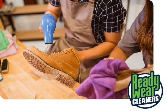 Two people use a hair dryer and a purple cloth to clean a tan suede boot at a workbench, with Ready Wear Cleaners logo.