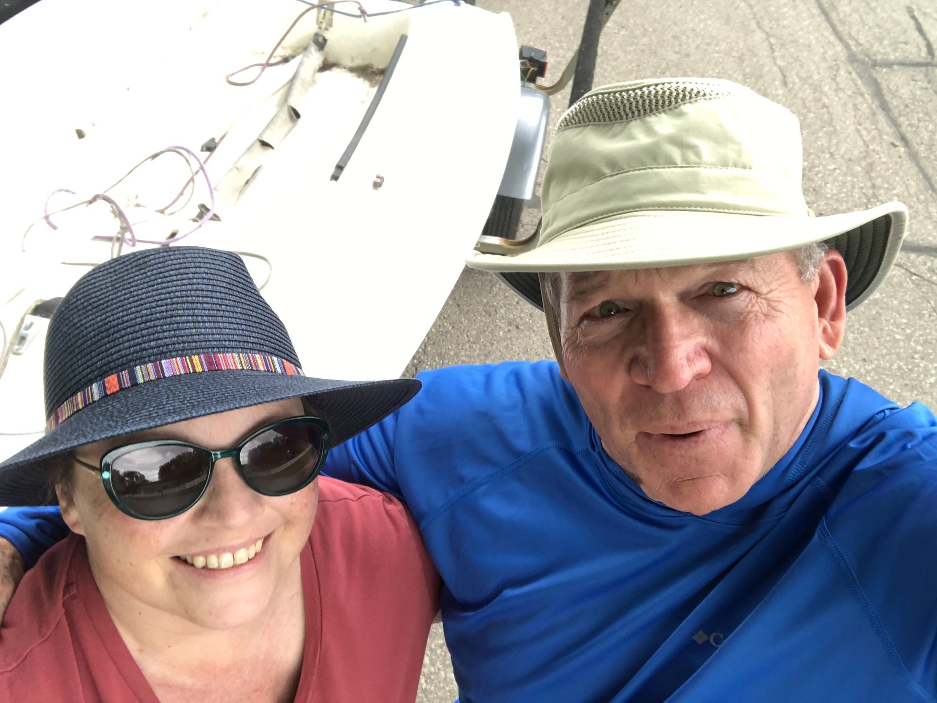Smiling couple in hats, next to a boat. The woman wears sunglasses, the man a blue shirt.