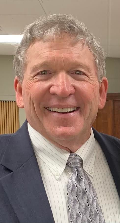 Three people in a meeting. Man in blazer gestures; two others seated, documents on table. Sunlight from window.